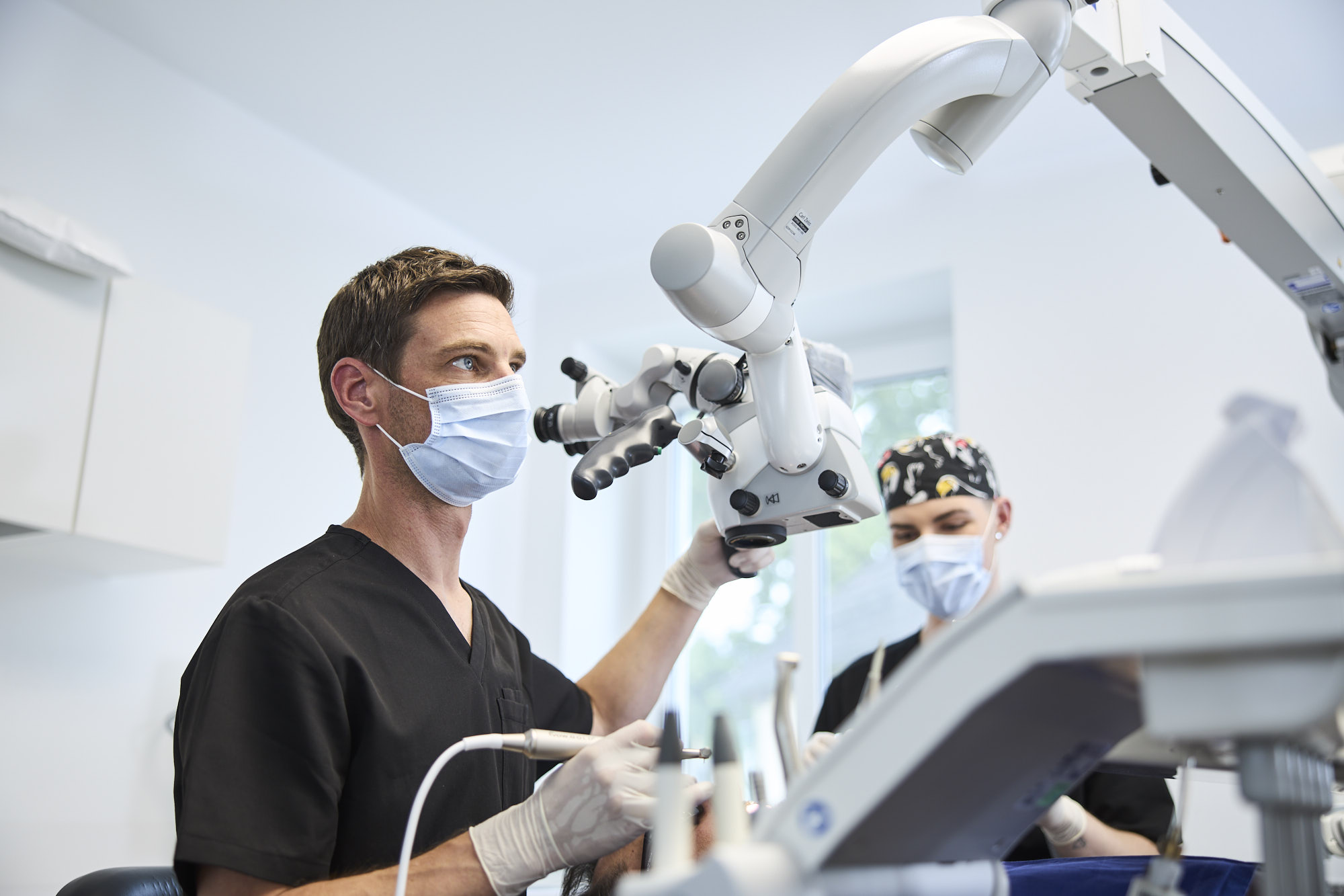 Der Zahnarzt hält sein Teleskop fest in der linken Hand und blickt vom Gerät weg nach vorne auf den Bildschirm. In der rechten Hand hält er ein Instrument. Im Hintergrund ist ein Behandlungsassistent zu sehen.