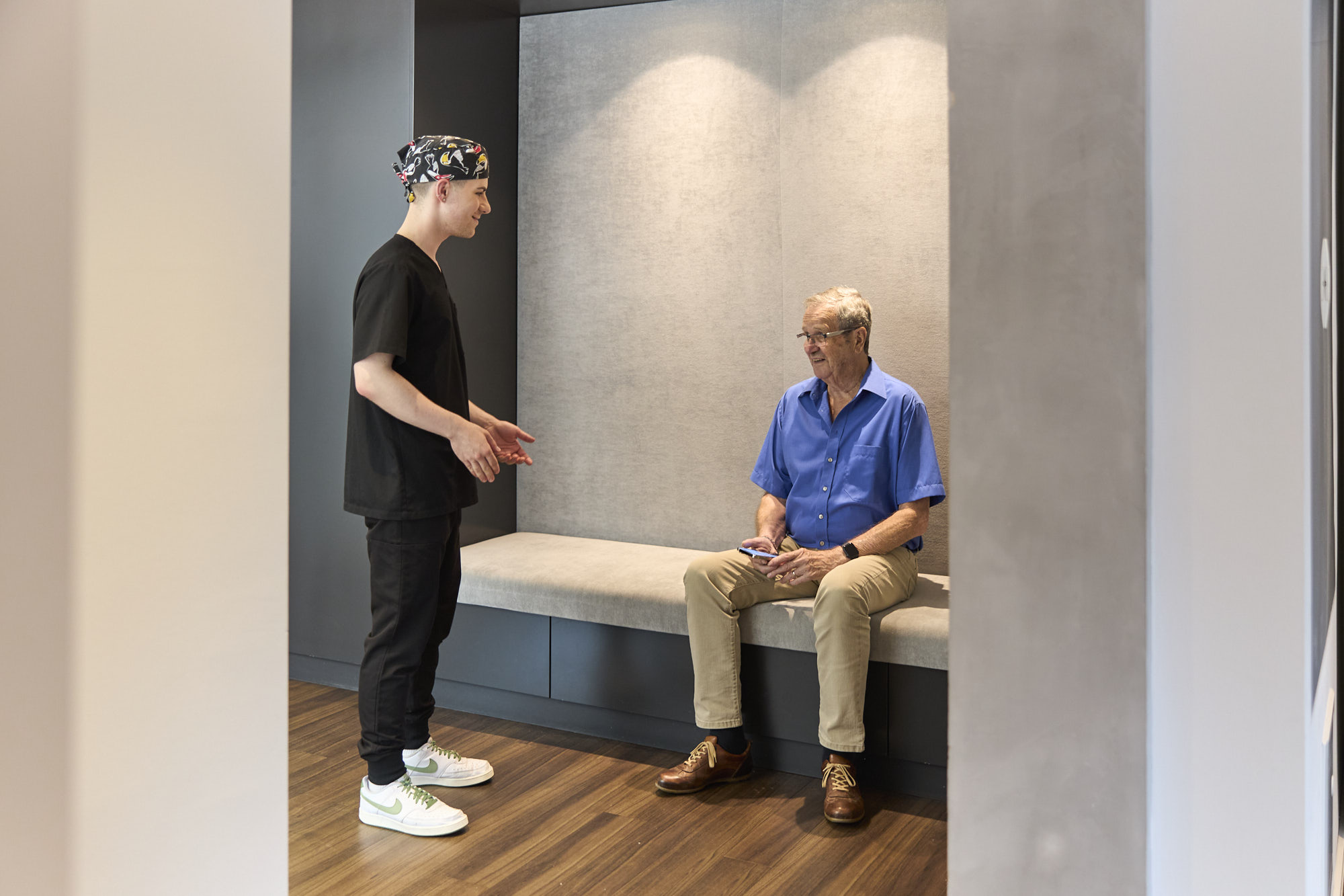 Ein älterer Patient sitzt im Wartebereich, während ein Behandlungsassistent vor ihm steht und erklärend mit den Händen gestikuliert.